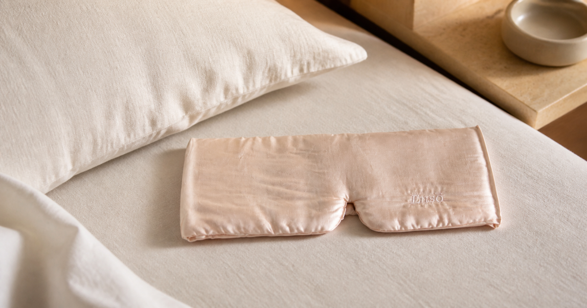 SILAIRA Ensō champagne silk sleep mask beside an ivory pillow in a warm neutral bedroom.